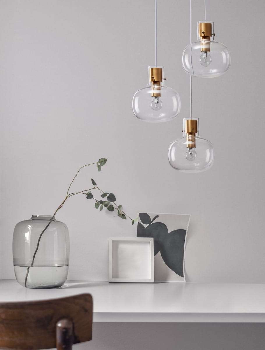 Lampa wisząca Padre z trzema kloszami, wykonana z przezroczystego szkła i złotych detalami. Klosze mają organiczny kształt, zawieszone na białych przewodach. W tle widoczny stół z wazonem, gałązką i ramką na zdjęcia.