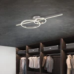 Lampa sufitowa Montilla z pilotem, w kolorze chromu, z trzema źródłami światła LED. Ułożona w nowoczesny sposób, z dwoma okrągłymi pierścieniami, znajdująca się na ciemnym suficie nad szafą z ubraniami.