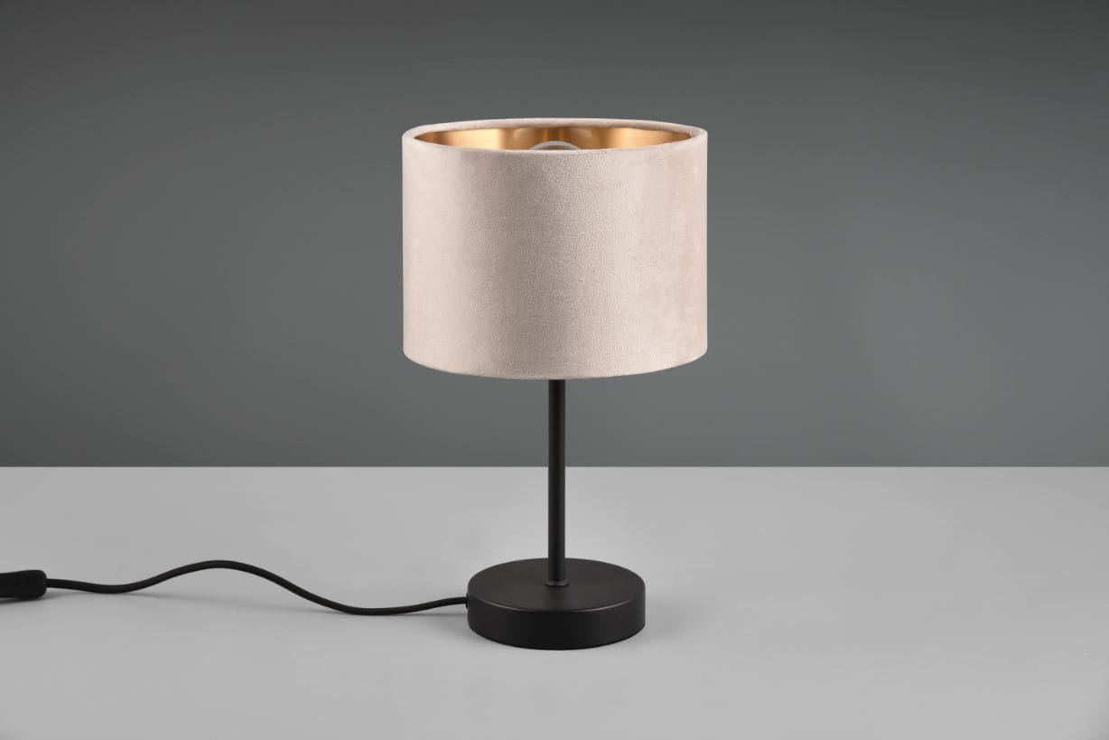 Lampa stołowa Julieta z czarną podstawą i jasnoróżowym kloszem, wykonana z materiału tekstylnego, z wykończeniem w złotym kolorze wewnątrz. Ujęcie z boku, na jasnym stole, widoczny kabel zasilający.