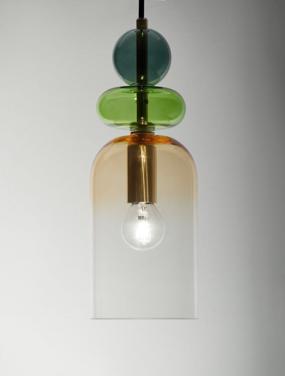 Lampa wisząca Balam 1xE14 w kolorze złotym z przezroczystym kloszem. Na górze znajdują się szklane elementy w kolorze zielonym i niebieskim. Klosz ma cylindryczny kształt, a żarówka jest dobrze widoczna wewnątrz. Produkt zawieszony na czarnym kablu.