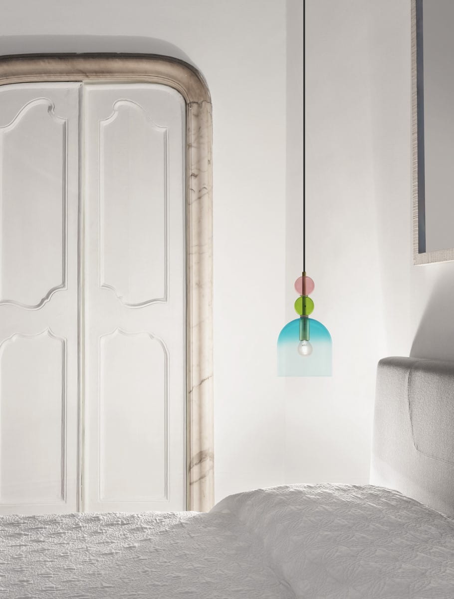 Lampa wisząca Balam 1xE14 w kolorze złotym z szklanym kloszem w odcieniach błękitu i zieleni. Posiada elegancką, minimalistyczną formę. Zawieszona na cienkim, czarnym przewodzie, w otoczeniu jasnych ścian i sypialnianego łóżka.