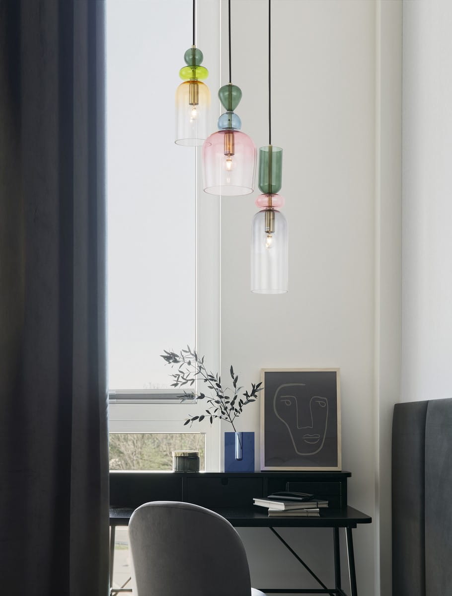 Lampa wisząca Balam 3xE14 w kolorze złotym, z trzema szklanymi kloszami w różnych kolorach: przezroczystym, różowym i zielonym, zawieszona na czarnych przewodach. Ujęcie z boku, w nowoczesnym wnętrzu z biurkiem i dekoracjami.