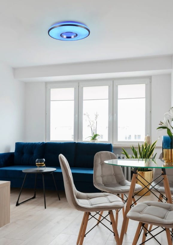Plafon Realta z pilotem, RGB, 1xLED/24W, wykonany z aluminium. Oświetlenie w niebieskim kolorze, zamontowane na suficie w jasnym wnętrzu z oknami, zielonymi roślinami, niebieską sofą i szklanym stołem.