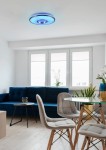 Plafon Realta z pilotem, RGB, 1xLED/24W, wykonany z aluminium. Oświetlenie w niebieskim kolorze, zamontowane na suficie w jasnym wnętrzu z oknami, zielonymi roślinami, niebieską sofą i szklanym stołem.