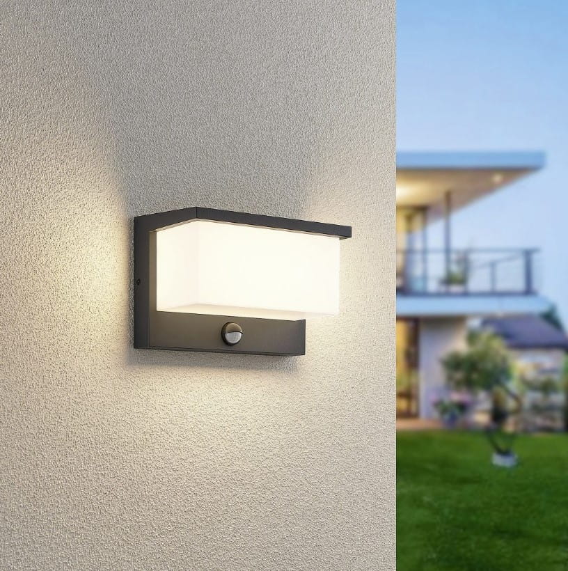 Kinkiet zewnętrzny Nestos LED w kolorze antracytowym, prostokątny, zamontowany na ścianie, z białym kloszem i czujnikiem ruchu. Produkt o nowoczesnym designie, umieszczony w aranżacji elewacyjnej domu, na tle zielonego trawnika.