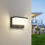 Kinkiet zewnętrzny Nestos LED w kolorze antracytowym, prostokątny, zamontowany na ścianie, z białym kloszem i czujnikiem ruchu. Produkt o nowoczesnym designie, umieszczony w aranżacji elewacyjnej domu, na tle zielonego trawnika.