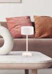 Lampa stołowa Jeff w kolorze białym z LED o mocy 1,5W. Ma cylindryczny klosz na cienkiej nogi i okrągłą podstawę. Ustawiona na stoliku, obok dekoracyjnego wazonu, w aranżacji z kanapą w odcieniach brązu i beżu.