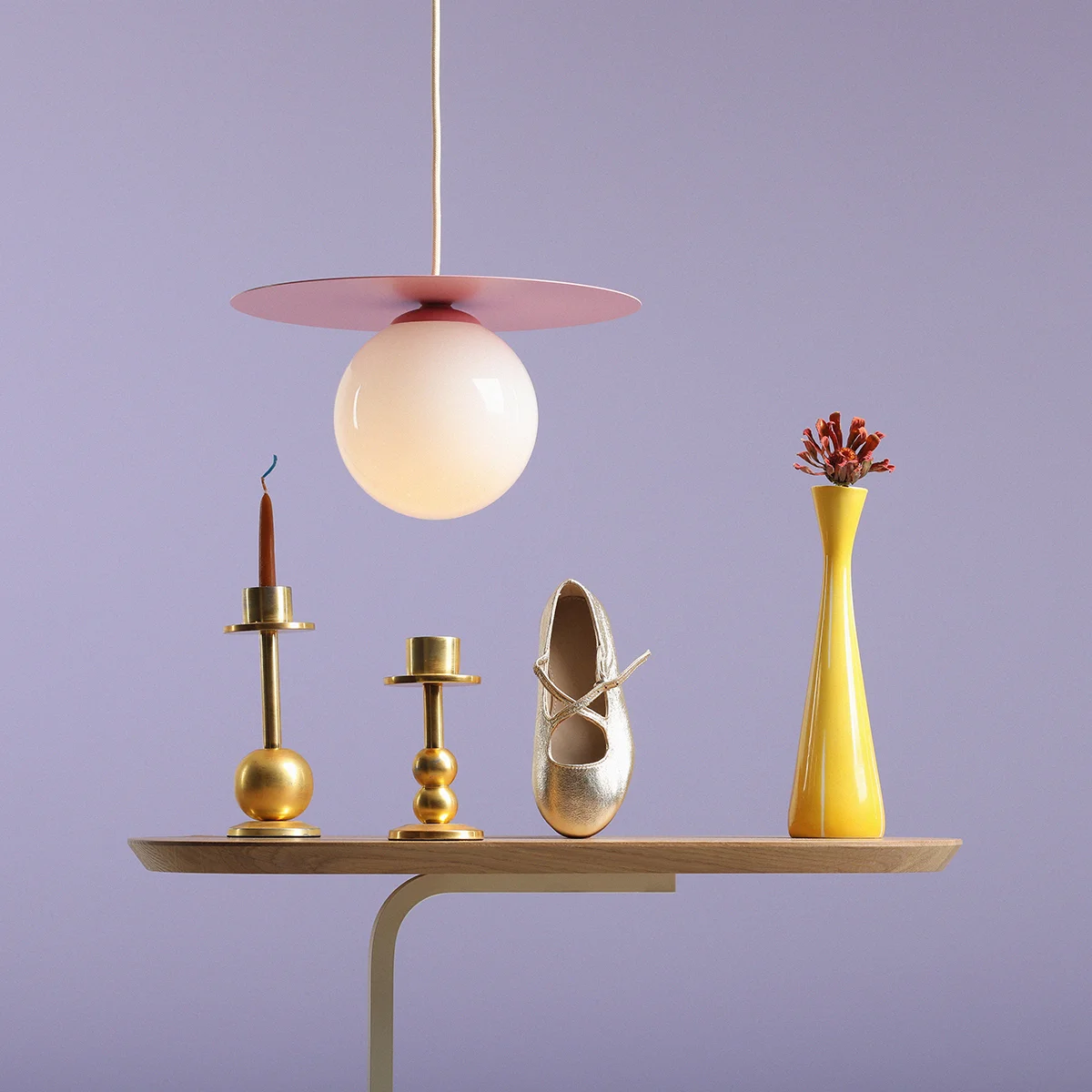 Lampa wisząca Loop Baby Pink S z okrągłym kloszem w kolorze białym i różową podstawą, zawieszona nad drewnianym blatem. W tle złote świeczniki, złota baletka i żółty wazon z kwiatami, na fioletowym tle.