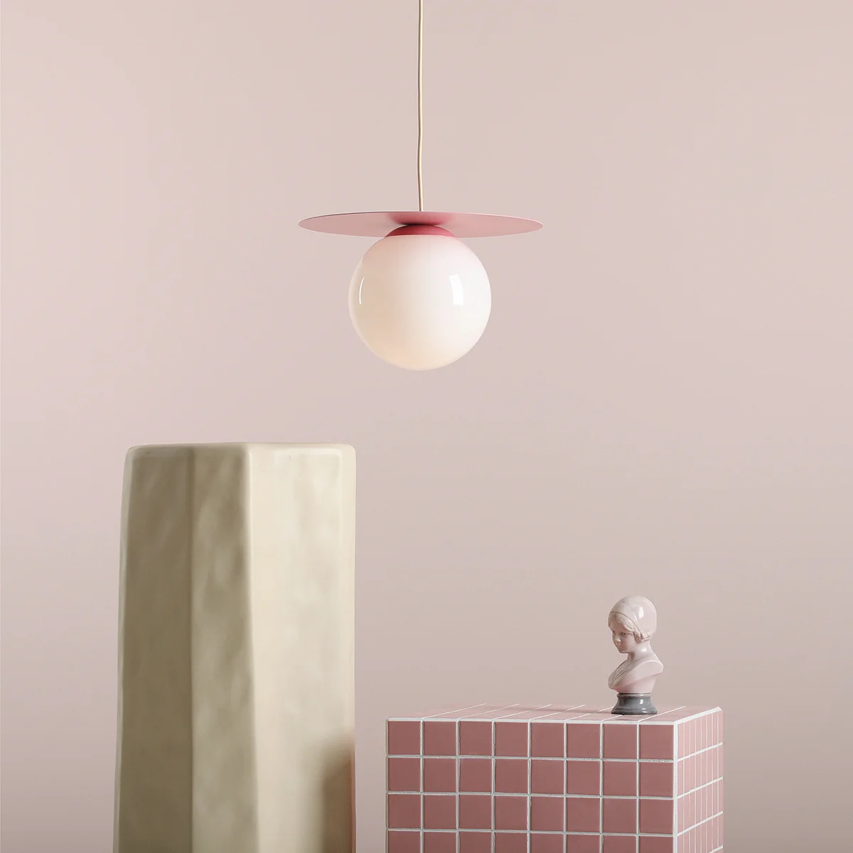 Lampa wisząca Loop Baby Pink M z kulistym kloszem w kolorze białym i różowym metalowym daszkiem. Zawieszona na cienkim przewodzie, w otoczeniu geometrycznych dekoracji w pastelowych odcieniach. Idealna do nowoczesnych wnętrz.