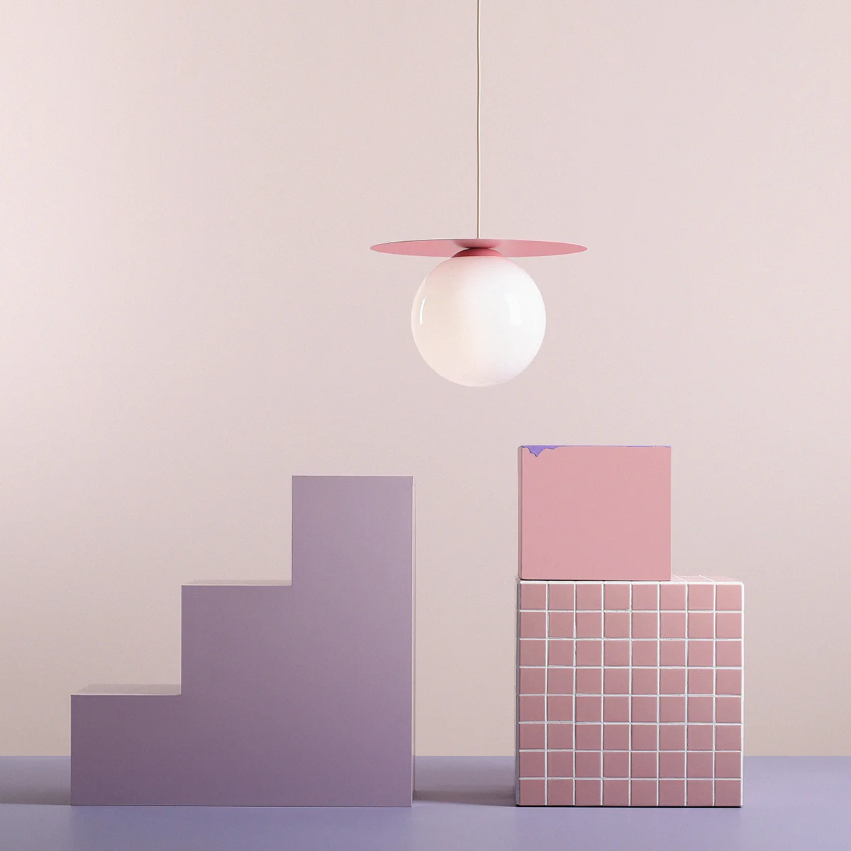 Lampa wisząca Loop Baby Pink L z kloszem w kształcie kuli, umieszczona na różowej podstawie. Produkt wisi nad geometrycznymi elementami w odcieniach fioletu i różu, na jasnym tle. Klosz wykonany z przezroczystego szkła, metalowe detale w kolorze różowym.