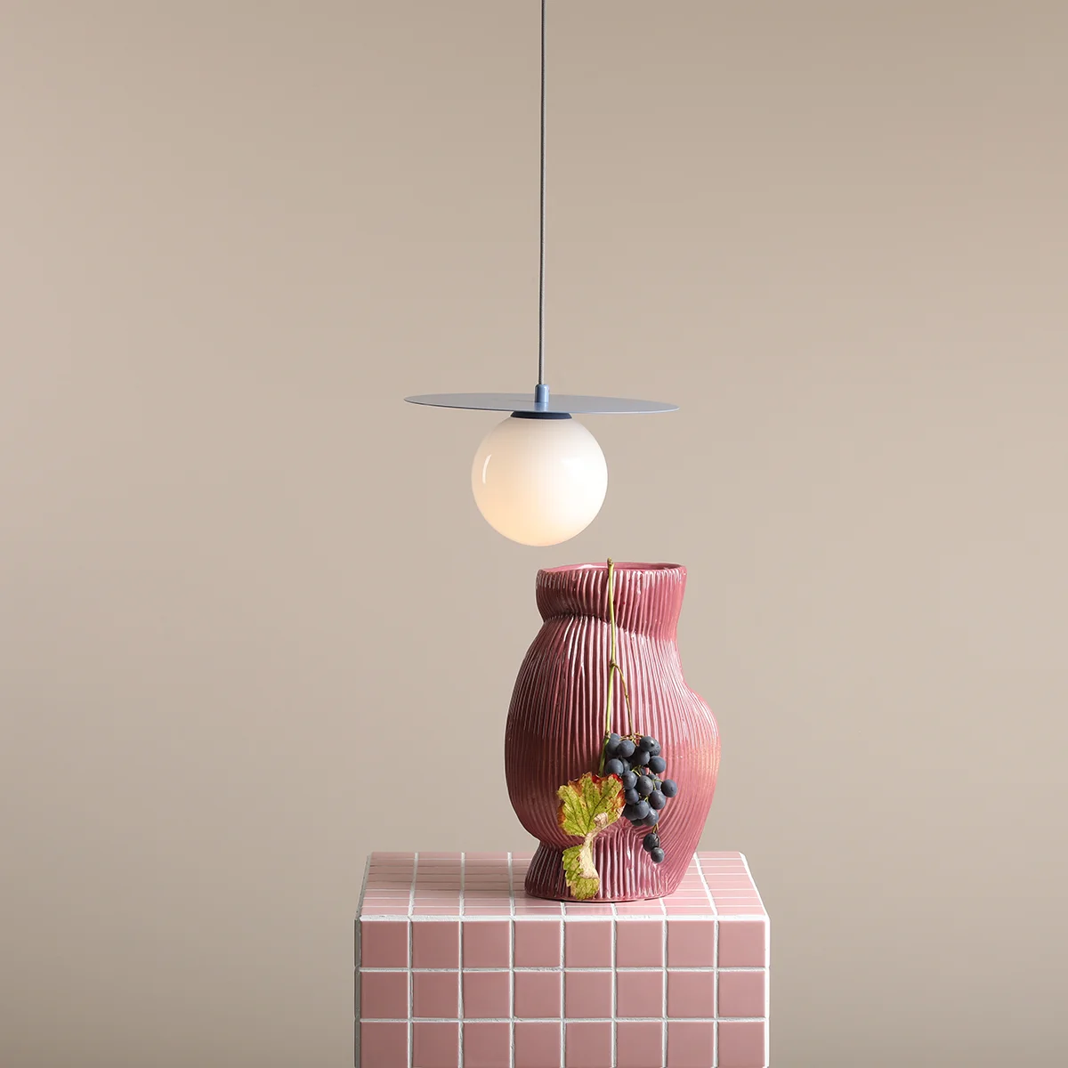 Lampa wisząca Loop Dusty Blue S z okrągłym kloszem z mlecznego szkła, zawieszona na cienkim przewodzie, nad ceramiczną wazonem w odcieniach różu z dekoracyjnymi winogronami. Tło w neutralnym kolorze.