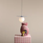 Lampa wisząca Loop Dusty Blue S z okrągłym kloszem z mlecznego szkła, zawieszona na cienkim przewodzie, nad ceramiczną wazonem w odcieniach różu z dekoracyjnymi winogronami. Tło w neutralnym kolorze.