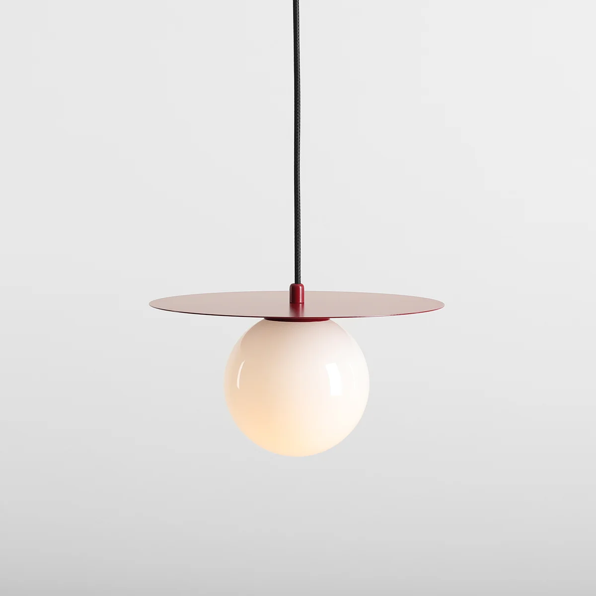 Lampa wisząca Loop Red Wine S z jednym źródłem światła E14. Posiada okrągłą, białą kulę i okrągłą, czerwoną osłonę na górze. Wisi na czarnym kablu, na jasnym tle.