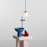Lampa wisząca Loop Red Wine z białym, okrągłym kloszem i czerwonym wykończeniem, zawieszona na czarnym przewodzie. W tle niebieska miska i zestaw czerwonych cylindrów na różowym stoliku. Prosty, minimalistyczny design na jasnym tle.