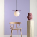 Lampa wisząca Loop Red Wine L z okrągłą, białą kulą, zawieszona na cienkim czarnym przewodzie. Posiada czerwony, płaski element na górze. W tle widoczna jasnofioletowa ściana, drewniane krzesło i rzeźbiony wazon na białym stoliku.