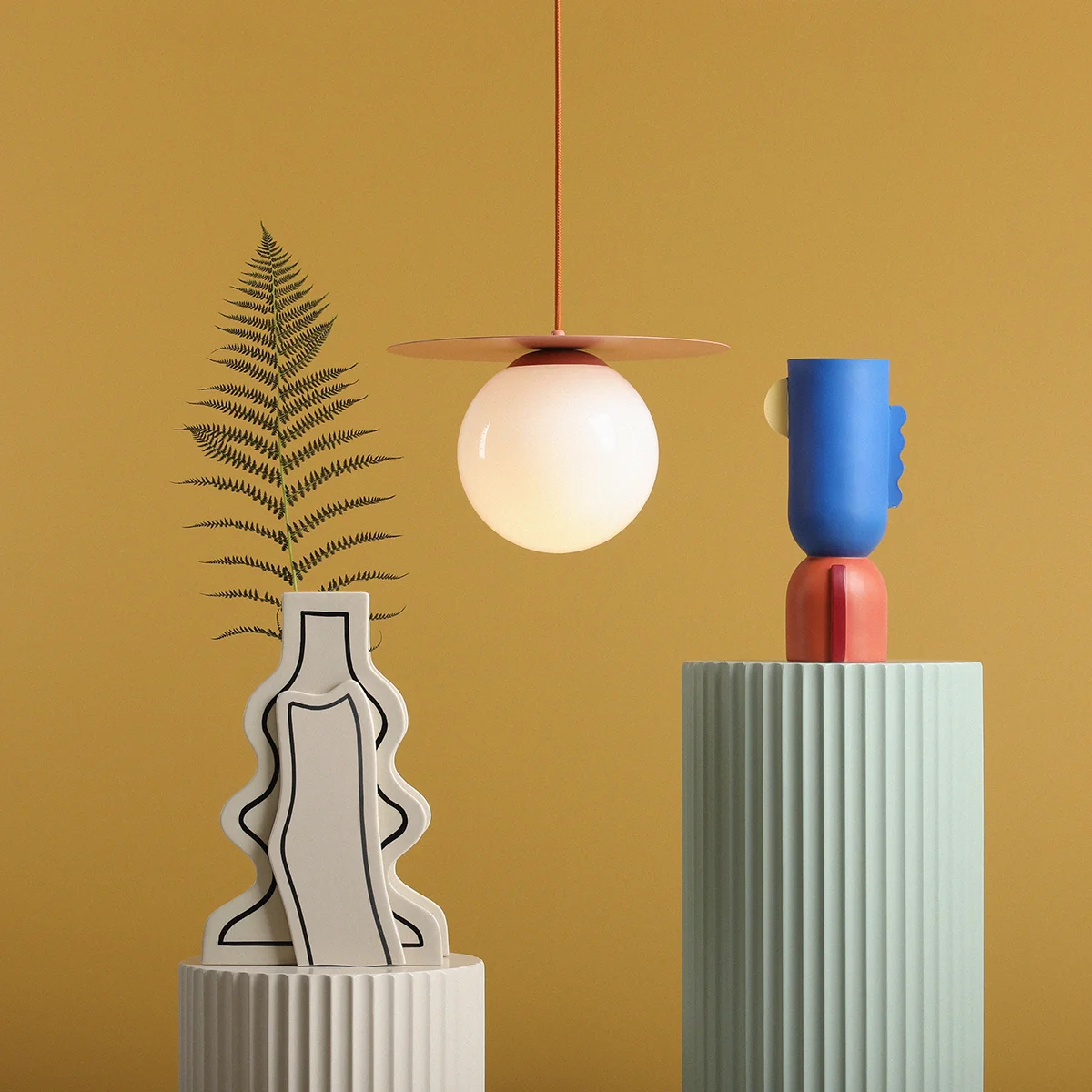 Lampa wisząca Loop Coral M z kloszem w kształcie kuli, zawieszona na pomarańczowym przewodzie. W tle dwa dekoracyjne wazony w pastelowych kolorach, z rośliną w jednym oraz abstrakcyjną figurą w drugim, na jednolitym żółtym tle.