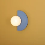 Kinkiet Loop Dusty Blue z okrągłą, białą kulą i półokrągłym, błękitnym elementem. Zamontowany na żółtej ścianie, prezentuje nowoczesny design. Kształt geometryczny podkreśla minimalistyczny styl, idealny do różnych aranżacji wnętrz.