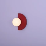 Kinkiet Loop Red Wine z jedną oprawą E14, składający się z półokrągłej, ciemnoczerwonej osłony i białej, okrągłej żarówki. Produkt zamontowany na jasnofioletowej ścianie, w pozycji stojącej, z widocznym detalem na gładkiej powierzchni.