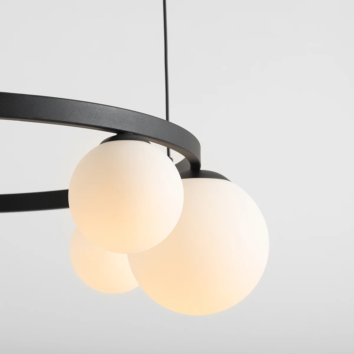 Lampa wisząca Garda w kolorze czarnym, z pięcioma okrągłymi kloszami z matowego szkła. Konstrukcja w kształcie pierścienia, zawieszona na cienkim kablu. Widać trzy klosze, każdy w odcieniu białym, emitujące ciepłe światło. Tło jasne i jednolite.