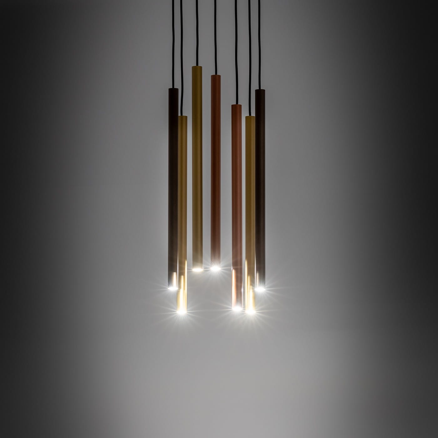Lampa wisząca 7-punktowa w kolorach mosiężnym, złotym i brązowym, z prostymi, cylindrycznymi kloszami. Ujęcie z dołu, prezentujące lampę zawieszoną na czarnych przewodach. Źródła światła emitują delikatne blaski. Tło w odcieniu szarości.