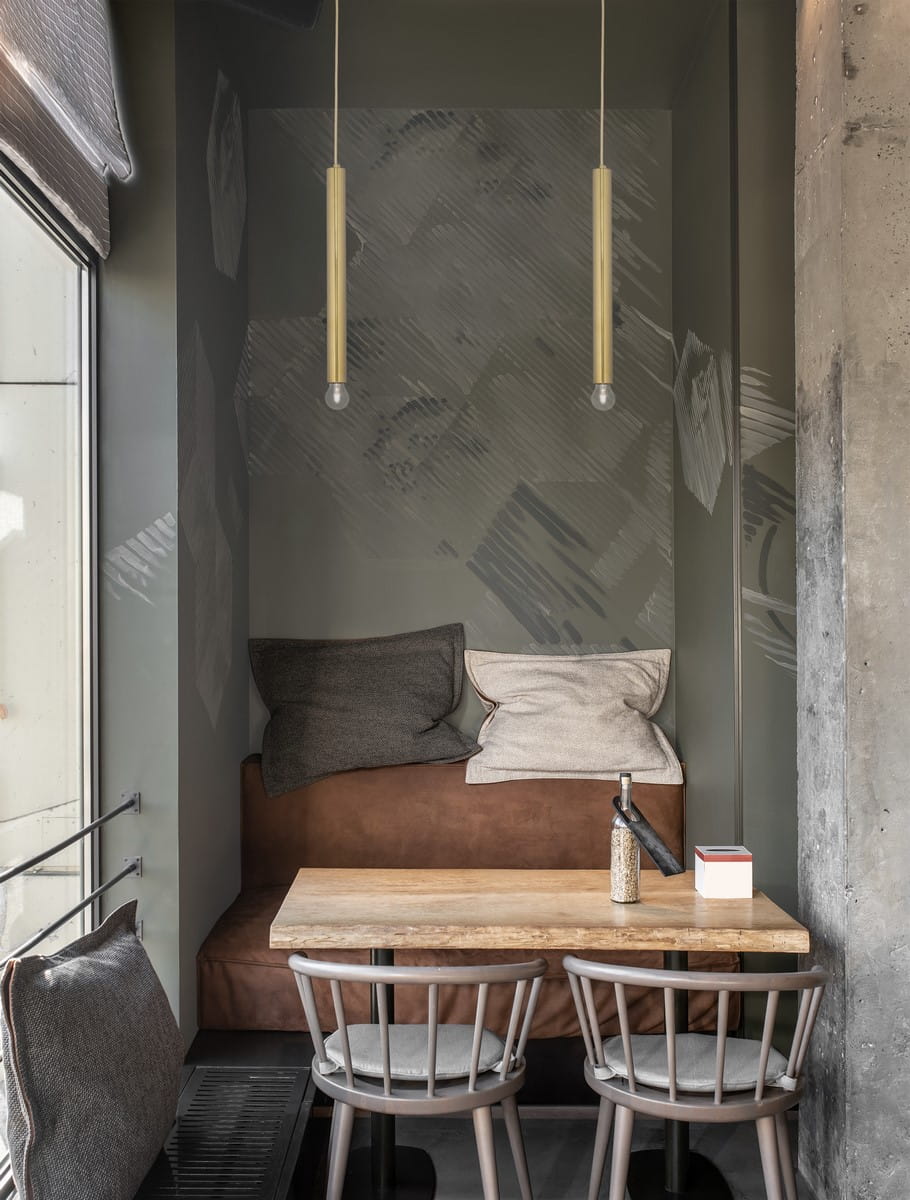 Wisząca lampa Bajan 35 z jedną oprawą E27 w złotym kolorze, umieszczona w nowoczesnym wnętrzu. Lampa ma prostą, cylindryczną formę i znajduje się nad drewnianym stołem w kąciku z miękkimi poduszkami. Tło w odcieniach szarości.