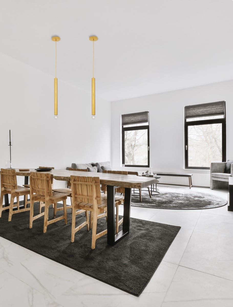 Lampa wisząca Bajan 50, złota, z jedną oprawą E27, zawieszona w nowoczesnym wnętrzu. Dwa cylindryczne klosze na długich przewodach, w minimalistycznym stylu, nad drewnianym stołem i krzesłami. Tło jasne, z oknami i meblami w stonowanej kolorystyce.