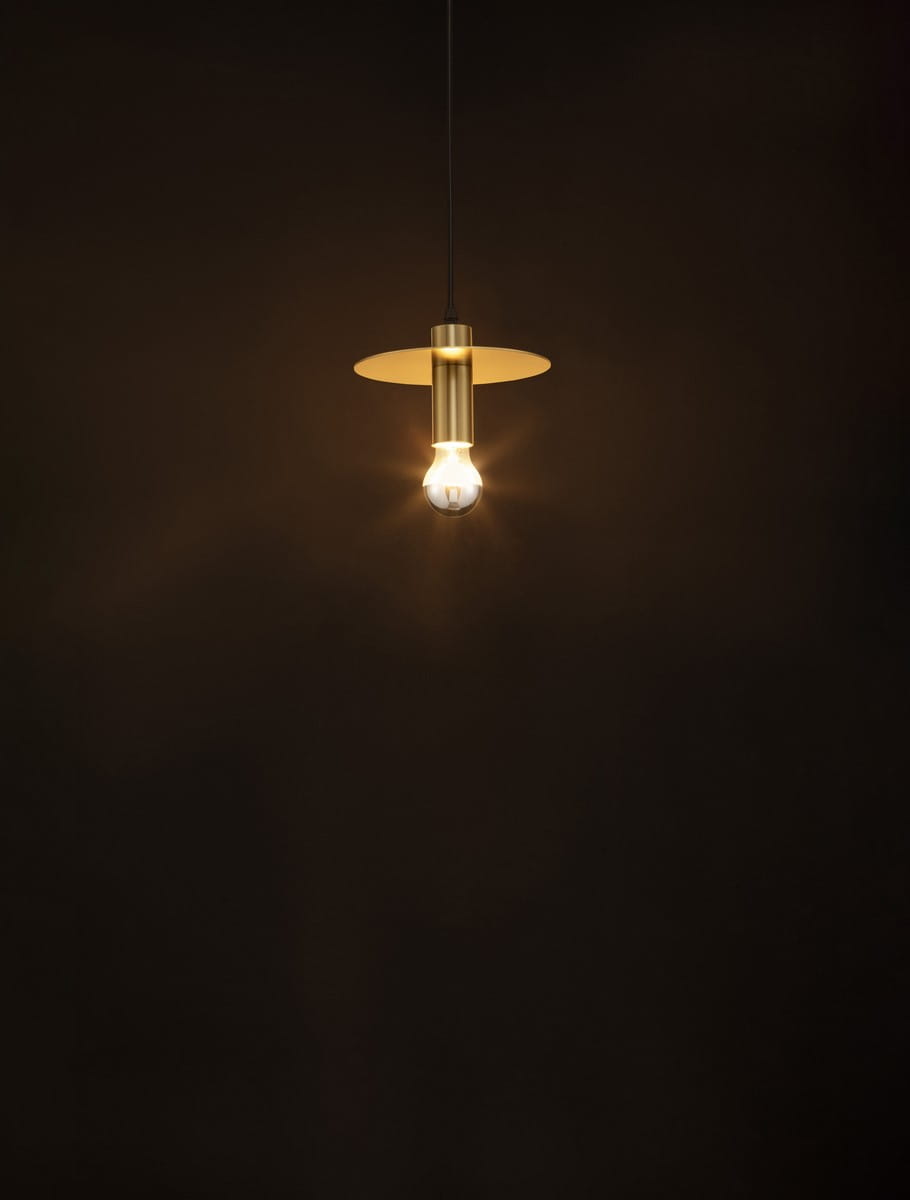 Lampa wisząca Bajio 20, kolor złoty, z jednym źródłem światła E27. Ujęcie frontalne, lampa zawieszona na czarnym kablu, z okrągłym kloszem nad żarówką, emitującym ciepłe światło na tle ciemnej ściany.