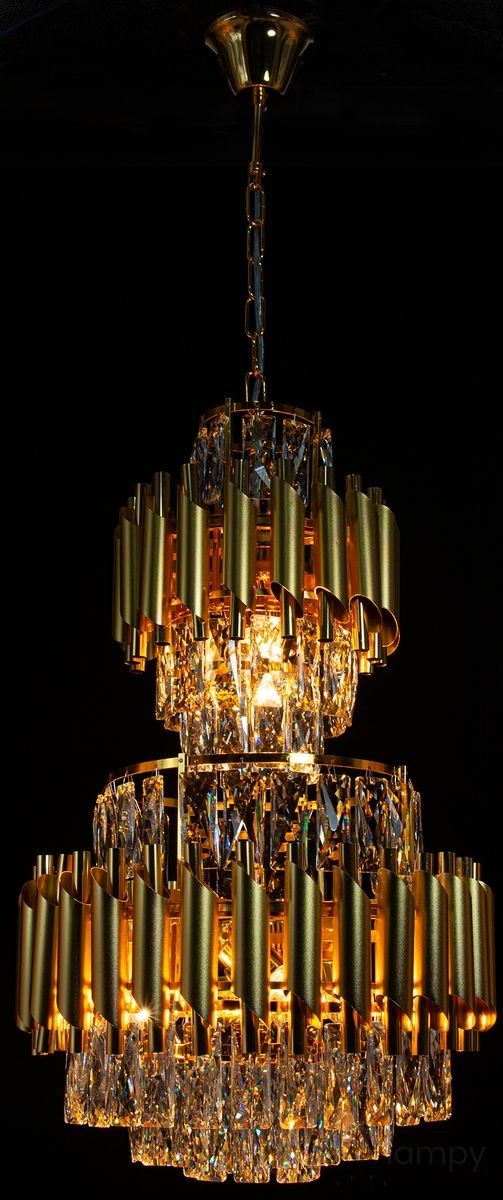Lampa wisząca Wersal z 6 gniazdami E27, w kolorze chromu i czerni. Posiada dwa poziomy dekoracyjnych, falistych elementów w złotym kolorze oraz kryształowe detale, które odbijają światło. Montowana na długim łańcuchu.