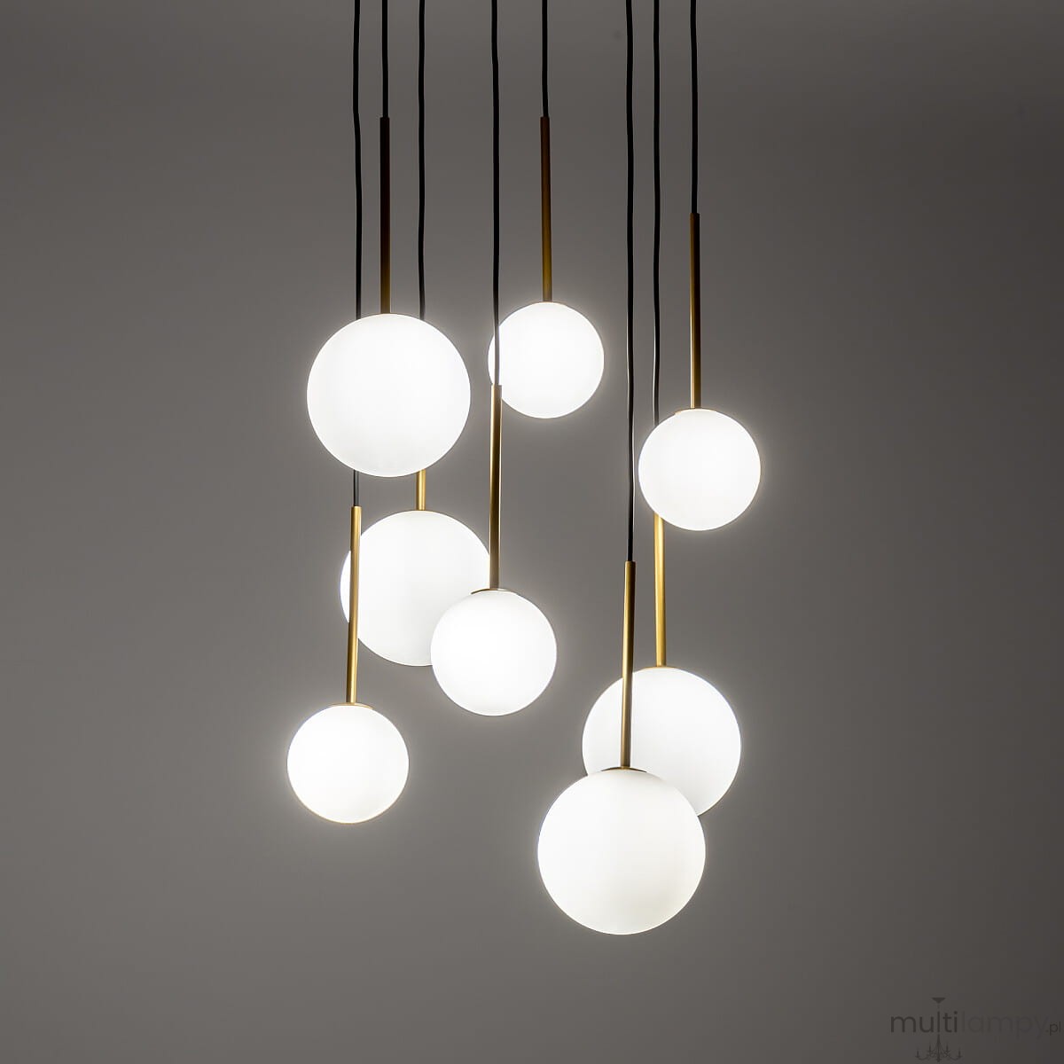 Lampa wisząca Karo z ośmioma kulistymi kloszami w kolorze białym, zawieszona na czarnych przewodach, z złotymi trzpieniami. Klosze różnej wielkości tworzą dynamiczny układ, na tle szarej ściany.