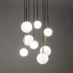 Lampa wisząca Karo z ośmioma kulistymi kloszami w kolorze białym, zawieszona na czarnych przewodach, z złotymi trzpieniami. Klosze różnej wielkości tworzą dynamiczny układ, na tle szarej ściany.