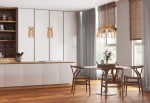 Lampa wisząca Kymi B w naturalnym kolorze, z widoczną konstrukcją z drewna, zawieszona nad okrągłym białym stołem. W tle nowoczesna kuchnia z szafkami i drewnianą podłogą. Dwa źródła światła w lampie, w stylu skandynawskim.