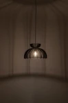 Czarna lampa wisząca Kymi C z kloszem w formie ażurowej konstrukcji, umieszczona w centralnej części na cienkim przewodzie. Widoczna żarówka E27, rzucająca cień na ścianę. Lampa wisi w prostym otoczeniu na jednolitym tle.