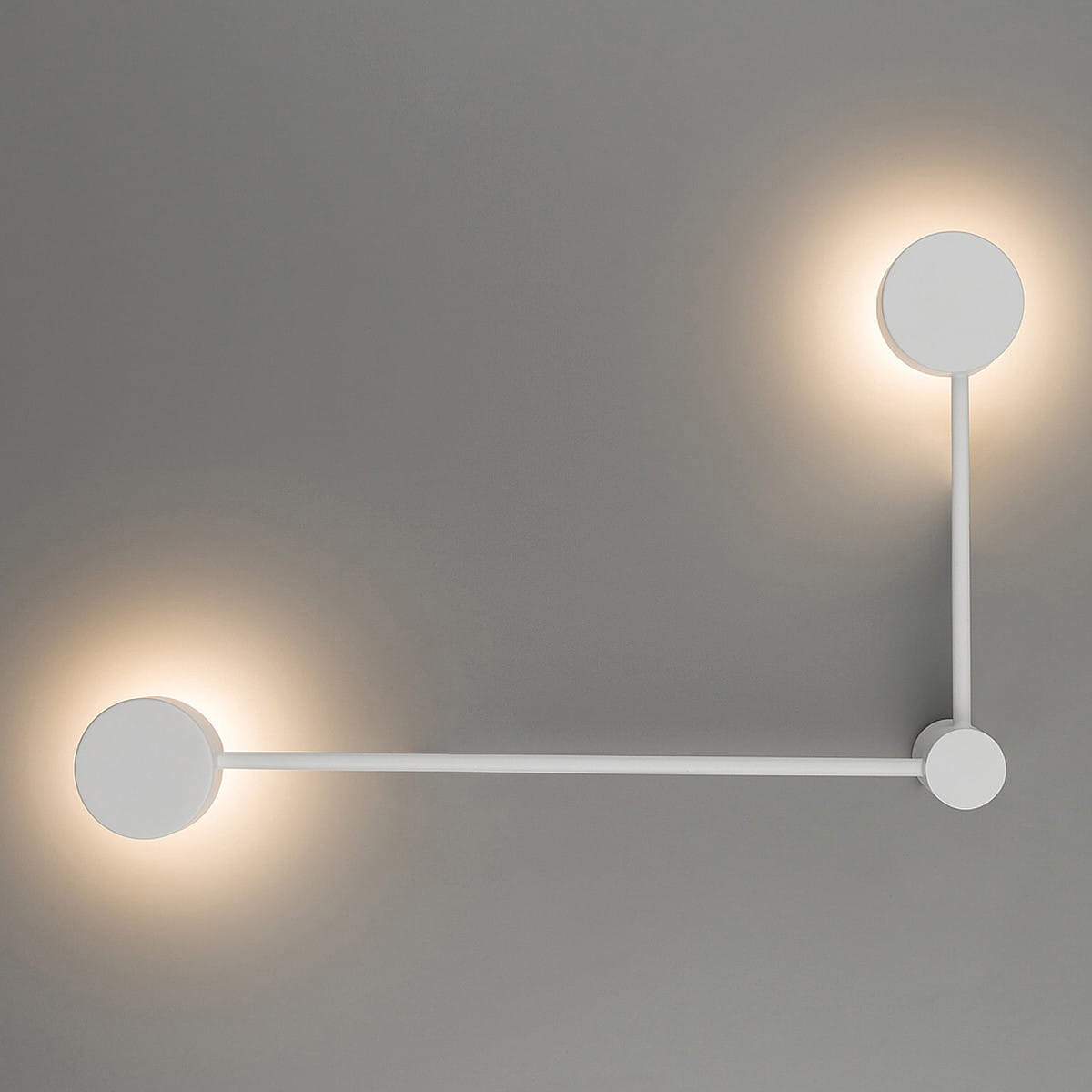 Biały kinkiet Orbit L z dwoma okrągłymi źródłami światła GX53, zamontowany na szarej ścianie. Produkt ma minimalistyczny design z prostym, poziomym ramieniem łączącym dwa światła, które emitują delikatne oświetlenie.