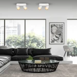 Lampa sufitowa Andy w kolorze białym i złotym, z dwoma uchwytami na żarówki GU10. Umieszczona na suficie, skierowana w dół, idealna do oświetlenia wnętrza nowoczesnego salonu. W tle widoczny czarny narożnik i okrągły stół z szklanym blatem.