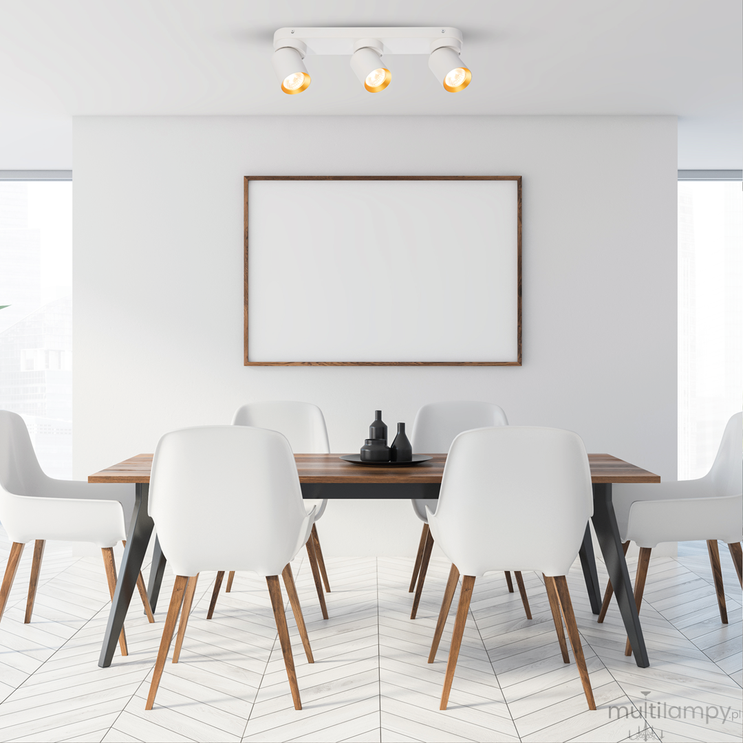 Biała lampa sufitowa typu spot z trzema oprawami GU10 w złotych detalach, zamontowana na suficie. W tle widoczna jasna aranżacja jadalni z drewnianym stołem i białymi krzesłami.