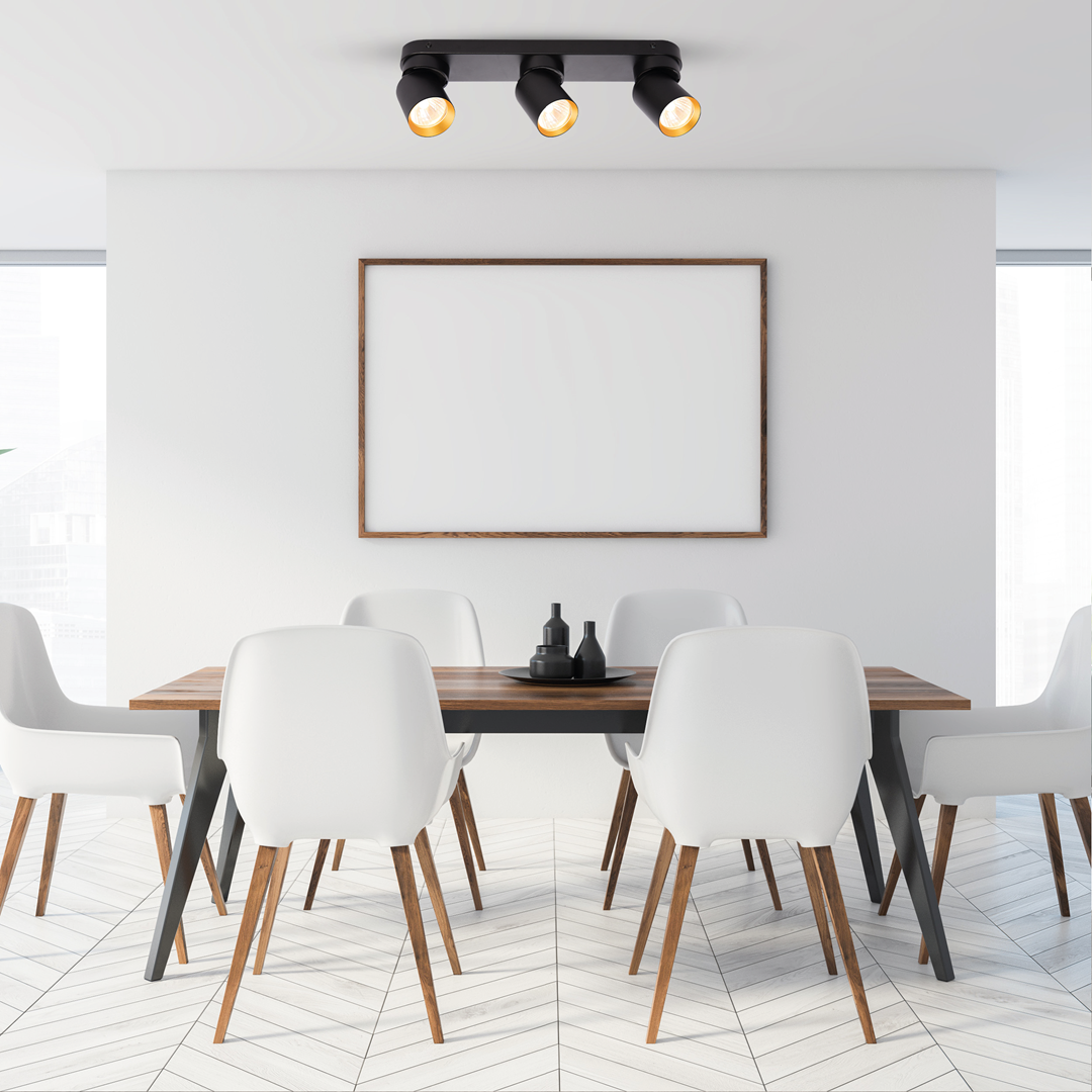 Czarna lampa sufitowa typu spot z trzema źródłami światła GU10, zamontowana na suficie w nowoczesnym wnętrzu. W tle widoczny stół z białymi krzesłami i dekoracjami na stole, na ścianie pusta ramka.