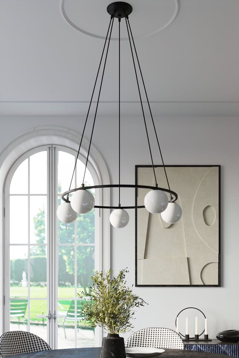 Lampa wisząca Yoli w kolorze czarnym z pięcioma źródłami światła G9. Okrągła konstrukcja z białymi kulistymi kloszami, zwisająca na cienkich linkach. Umieszczona w jasnym wnętrzu z dużym oknem, na tle minimalistycznych dekoracji i stołu.
