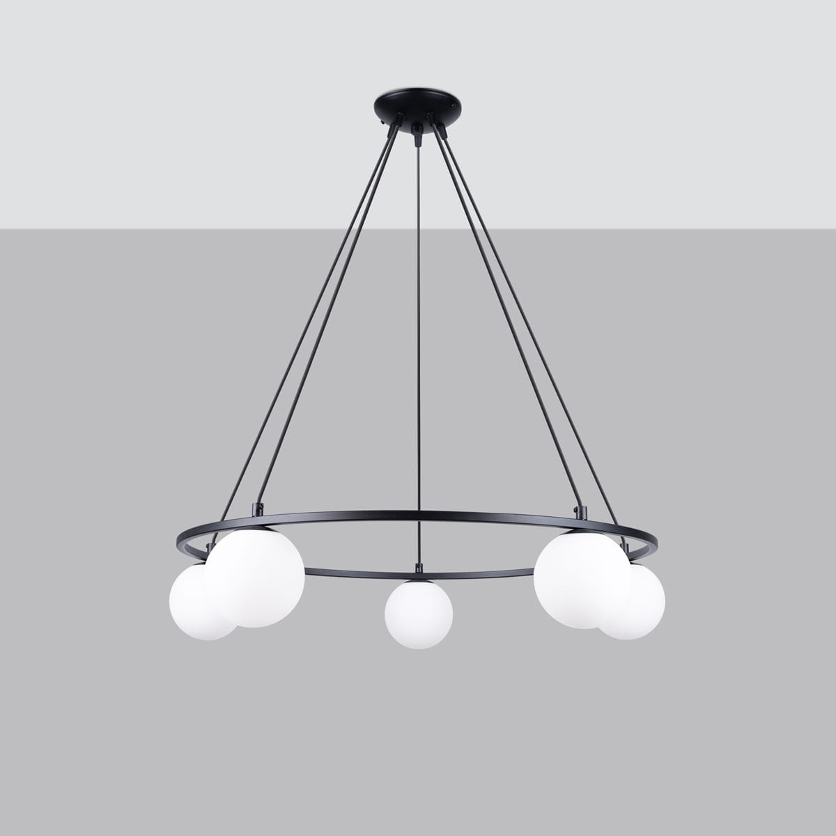 Lampa wisząca Yoli z pięcioma oprawami G9, czarna. Okrągła konstrukcja z metalowym pierścieniem i białymi, kulistymi kloszami, zawieszona na cienkich linkach. Tło w odcieniu szarości podkreśla minimalistyczny design lampy.