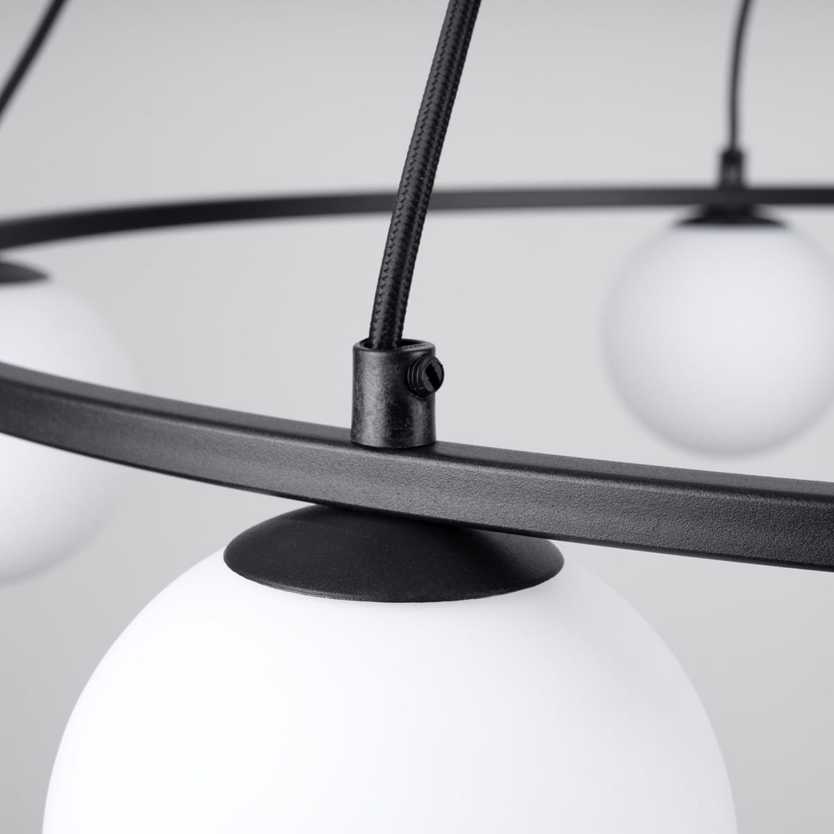 Lampa wisząca Yoli 5xG9 w kolorze czarnym, z metalowym pierścieniem i pięcioma białymi, okrągłymi kloszami. Widoczny detal łączenia kabla z oprawą. Produkt zwisający, w zbliżeniu na elementy konstrukcyjne.