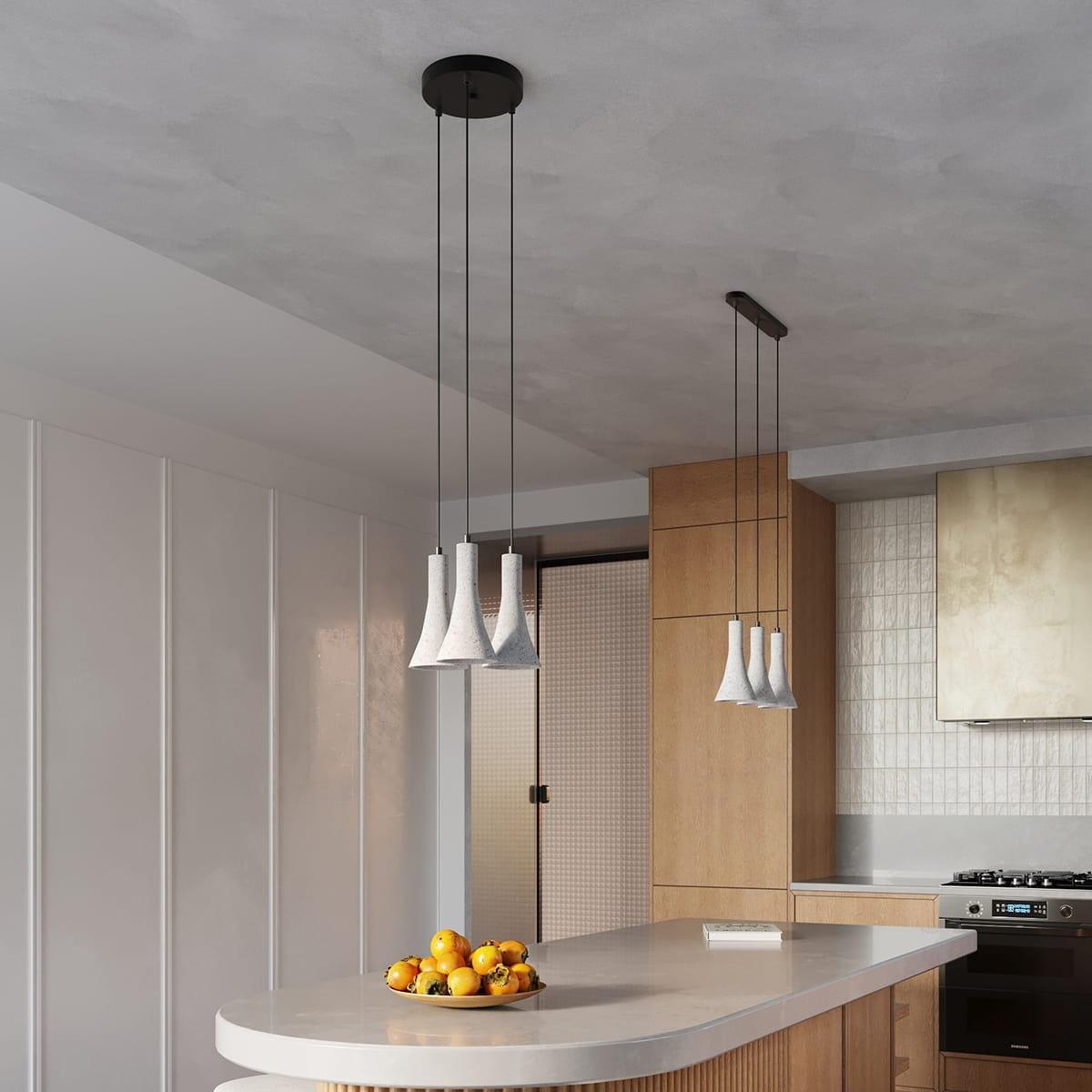 Lampa wisząca Rea w kolorze czarnym i betonowym z trzema kloszami, zawieszona na długich kablach. W tle widoczna wyspa kuchenna z owocami w misce oraz drewniane szafki. Oświetlenie idealne do nowoczesnych wnętrz.