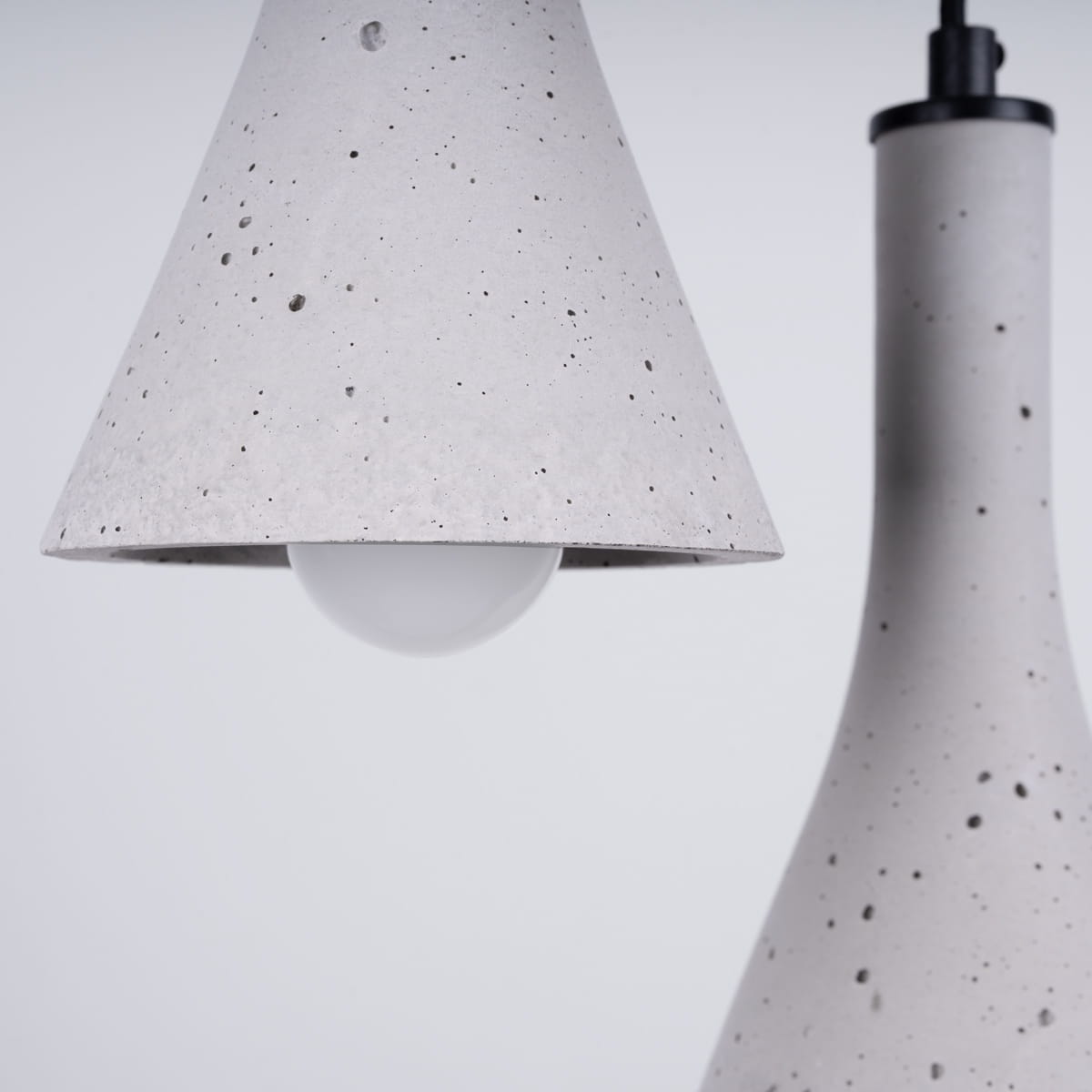 Lampa wisząca Rea w kolorze czarnym i betonowym, z trzema oprawkami E14. Widoczny klosz w kształcie stożka z szarego betonu z białą żarówką oraz smukły, betonowy element. Produkt zawieszony w pozycji pionowej na czarnym przewodzie.