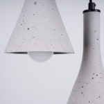Lampa wisząca Rea w kolorze czarnym i betonowym, z trzema oprawkami E14. Widoczny klosz w kształcie stożka z szarego betonu z białą żarówką oraz smukły, betonowy element. Produkt zawieszony w pozycji pionowej na czarnym przewodzie.