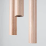 Lampa wisząca Pastelo z trzema tubami w kolorze drewna, wykonana z jasnego materiału. Ujęcie z bliska, ukazujące detale konstrukcji, z widocznymi otworami na źródła światła G9. Tło neutralne, szare.