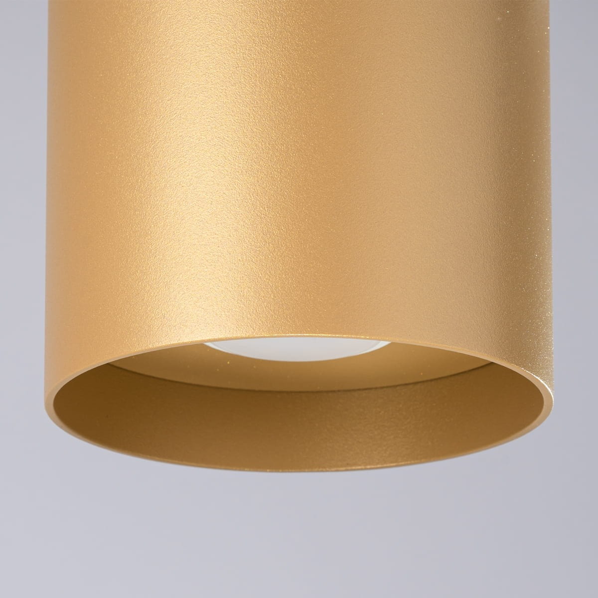 Lampa sufitowa Mika w kolorze złotym, o cylindrycznym kształcie, z miejscem na żarówkę GU10. Widoczny jest matowy metaliczny wykończenie, zbliżenie na górną część lampy, bez dodatkowych dekoracji.
