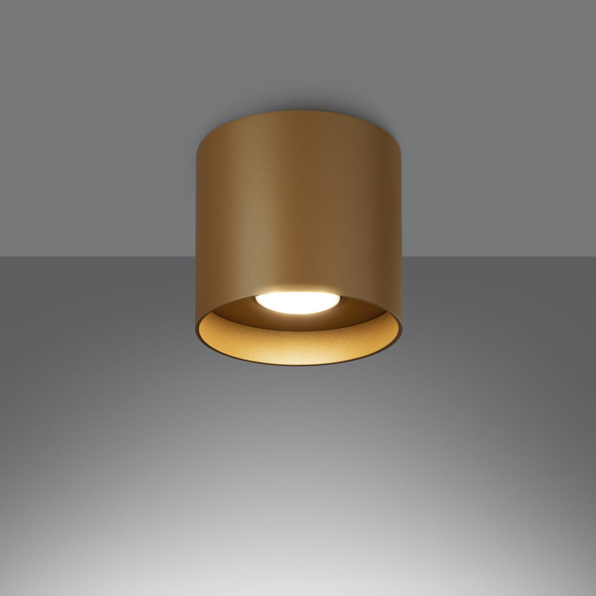 Lampa sufitowa Mika w kolorze złotym, o cylindrycznym kształcie, z widoczną oprawą na żarówkę GU10. Ujęcie frontalne, montowana na suficie, z białym wnętrzem dla lepszego odbicia światła. Tło szare, minimalistyczne.