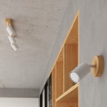 Kinkiet Verdo 1xGU10 w kolorze białym z jasnym drewnem, zamontowany na ścianie. Okrągła podstawa w jasnym drewnie, ruchoma lampa o cylindrycznym kształcie. Produkt umieszczony w nowoczesnym wnętrzu z drewnianymi półkami.