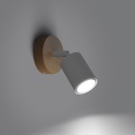 Kinkiet Verdo z białym kloszem w kształcie cylindrycznym, zamontowany na okrągłej podstawie z jasnego drewna. Ujęcie z boku, pokazujące źródło światła skierowane w dół. Tło w odcieniach szarości, podkreślające prostą formę lampy.