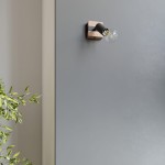 Kinkiet Zuca w kolorze czarnym z jasnym drewnem, zamontowany na szarej ścianie. Lampa z oprawą E27, z widoczną przezroczystą żarówką, umieszczona w narożniku. W tle delikatne liście rośliny.