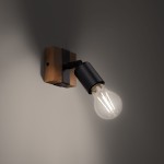 Kinkiet Zuca w kolorze czarnym z jasnym drewnem, z pojedynczym gniazdem E27. Lampa zamocowana na prostokątnej podstawie z drewna, z ruchomym ramieniem i żarówką w kształcie bańki, skierowaną w dół. Tło w odcieniu szaro-beżowym.