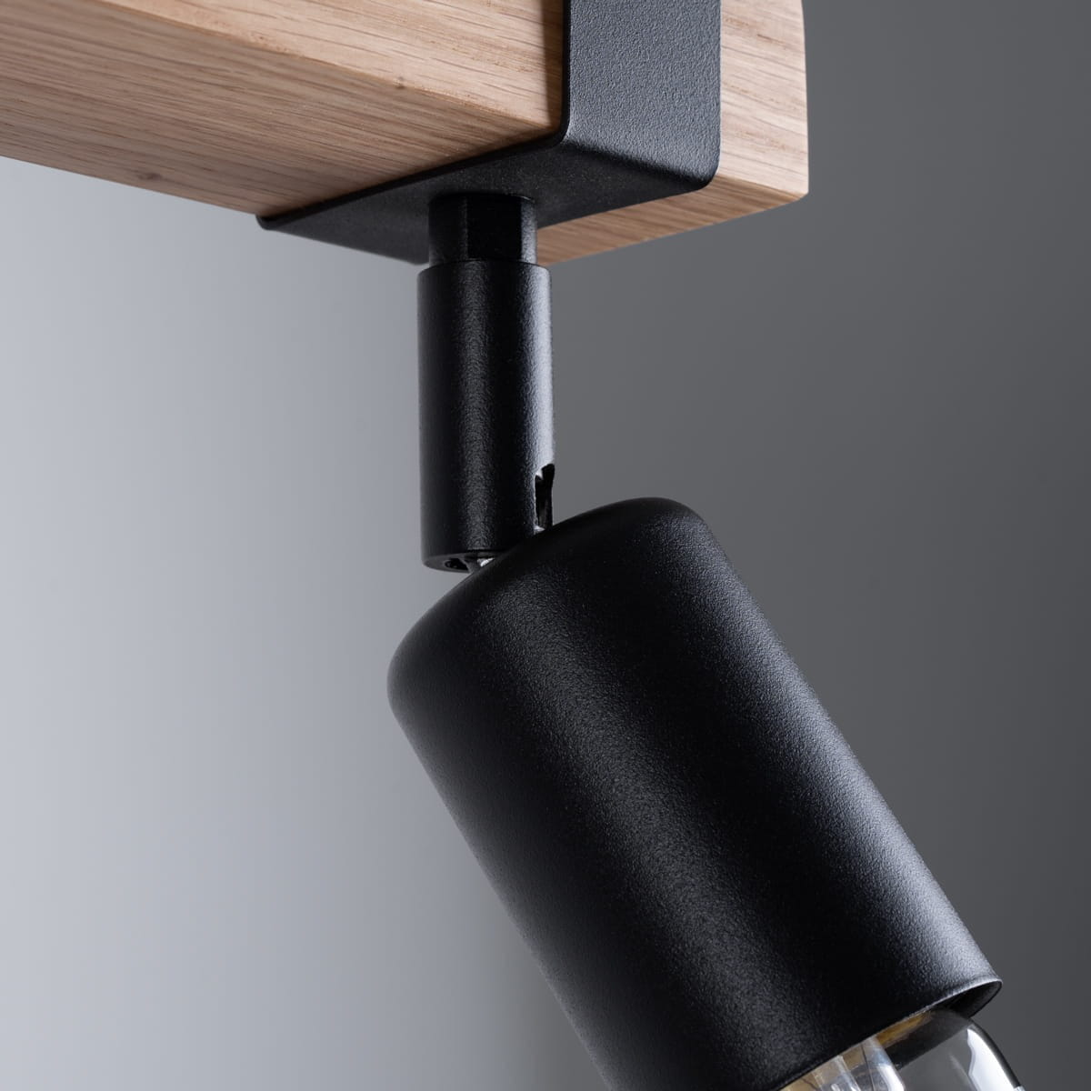 Lampa sufitowa Zuca z dwoma reflektorami E27, w kolorze czarnym z jasnym drewnem. Ujęcie z boku, ukazujące metalowy uchwyt i lampę w kształcie walca, zamontowaną na drewnianym stelażu. Tło w odcieniach szarości.